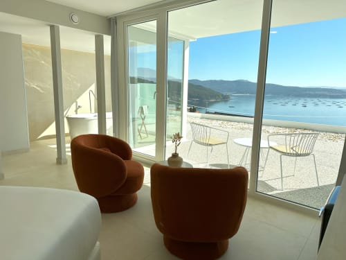 Habitación moderna del hotel Otantus en Muros con sillas burgundy curvas, vistas al océano y terraza de playa arenosa.