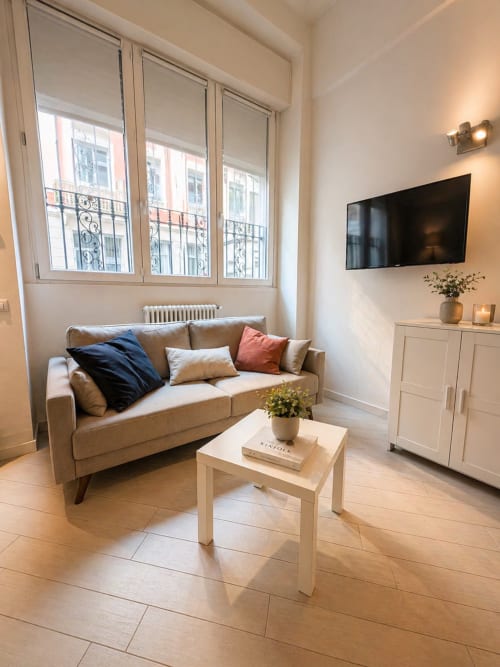 Modern loft living room in Milano with beige sofa, wooden table, TV, and Parisian-style windows with city view