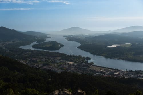 Vista aérea de Caminha, Portugal com rio sinuoso, colinas florestadas e montanhas distantes sob céu azul