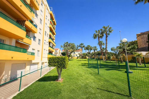 Edificio residencial amarillo y blanco con jardines paisajísticos verdes, palmeras y zona ajardinada vallada en Málaga, España