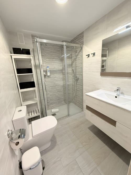 Baño moderno con ducha de acceso directo, lavamanos blanco, inodoro y estanterías de almacenamiento en apartamento Amapola Premium de Algarrobo Beach