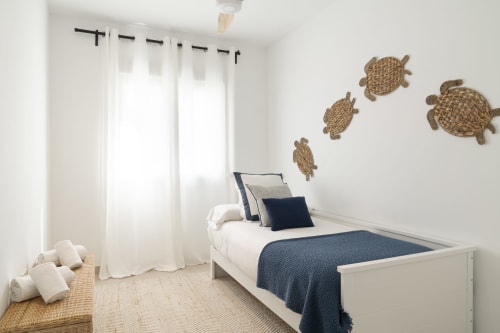 Dormitorio moderno con cama blanca, ropa de cama azul marino, decoración de tortuga tejida y cortinas translúcidas en el apartamento Axarquía Paraíso