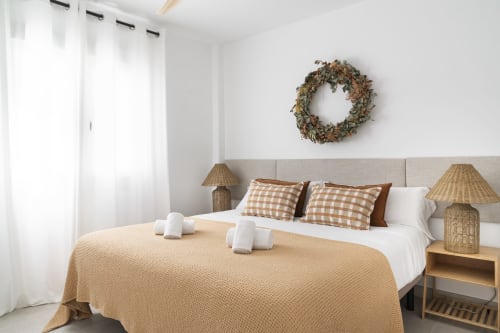 Luminoso dormitorio en el apartamento Axarquia Paraiso de Málaga con ropa de cama beige, lámparas de mimbre y cojines decorativos cuadrados