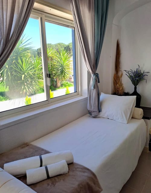 Quarto luminoso na Herdade da Cova da Onça com vista para o jardim, roupa de cama branca, toalhas enroladas e palmeiras visíveis