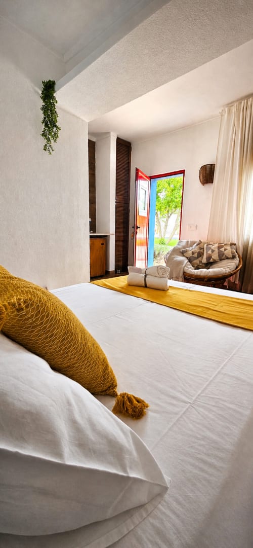 Quarto luminoso na Herdade da Cova da Onça com cama branca, detalhes em mostarda, móveis de madeira, porta com vista para jardim