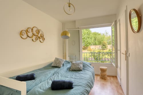 Chambre lumineuse avec literie bleue, art mural doré, luminaires suspendus et balcon vue jardin au Versailles Luxury Retreat