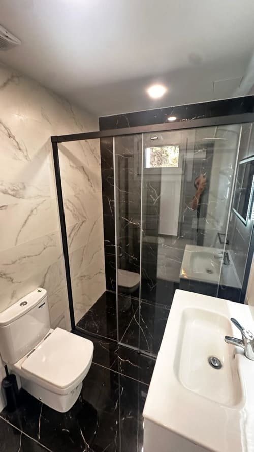 Baño moderno con paredes de mármol negro, lavabo blanco, inodoro y ducha con mamparas de cristal en Voramar Apartaments