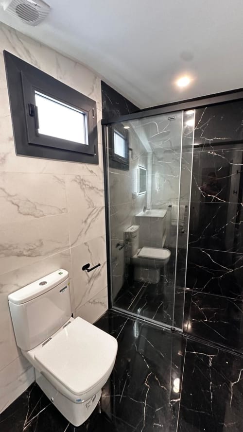 Baño moderno con azulejos de mármol negro, inodoro blanco, ducha de cristal y televisor de pared en villa Apartaments Voramar