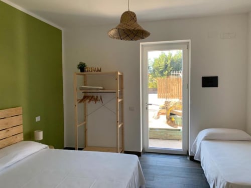 Camera tripla luminosa presso Cilento Rooms con due letti, parete verde, armadio in legno, lampada a sospensione intrecciata e porta in vetro