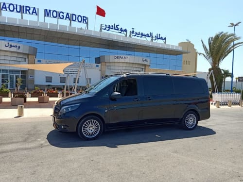 Monospace foncé garé au terminal de l'aéroport Mogador à Marrakech avec palmier et drapeau rouge visibles