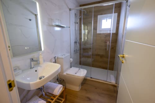 Baño moderno del Hotel Boutique Alcazaba Salobreña con lavamanos blanco, inodoro y ducha con cristal y acabado madera