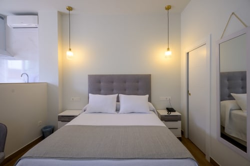 Dormitorio moderno de estudio con cabecera tapizada gris, ropa de cama blanca, lámparas colgantes, espejo y kitchenette