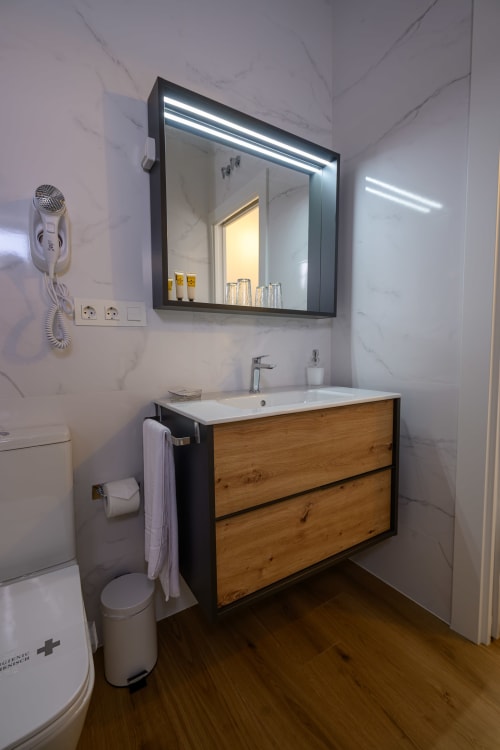 Baño moderno en Hotel Boutique Alcazaba Salobreña con mueble de madera, espejo LED, paredes de mármol blanco y servicios completos