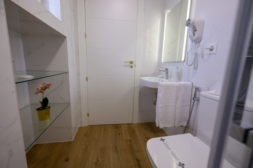 Baño moderno del hotel Alcazaba Salobreña con revestimiento blanco, piso de madera, lavamanos, inodoro y espejo fijado a la pared