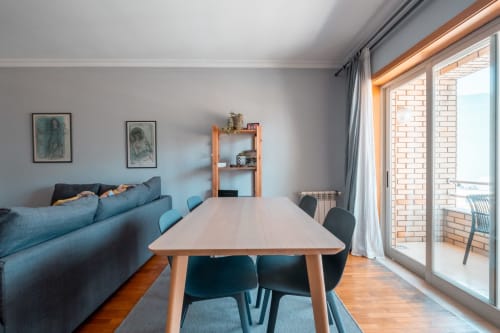 Área social integrada moderna no OceanSide EcoHost em Perafita com mesa de refeições, sofá teal, prateleiras de madeira e vistas citadinas