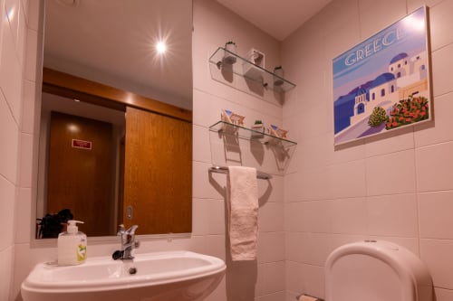 Banheiro moderno rosa com pia, vaso sanitário, toalheiro aquecido e pôster de viagens da Grécia no apartamento EcoHost em Perafita,