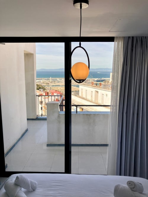 Chambre moderne à Tanger avec balcon vue mer, lampe suspendue et paysage côtier urbain visible par la baie vitrée