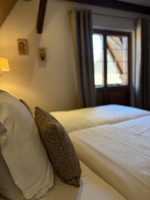 Chambre accueillante aux lits jumeaux à l'Hôtel de l'Ange à Niedermorschwihr avec literie crème, oreillers gris et portes ouvertes sur la terrasse