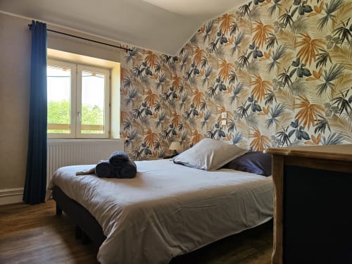 Chambre lumineuse aux Gîtes Les Muriers avec papier peint motifs, lit en bois, rideaux bleus et vue sur le jardin