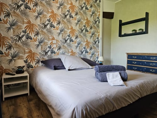 Chambre aux Gîtes Les Muriers avec papier peint motifs, lit blanc aux oreillers bleu marine, commode foncée et étagère murale noire