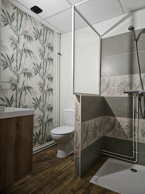 Salle de bain moderne avec papier peint motif feuilles de bambou, douche spacieuse, toilettes et meuble vasque en bois aux Gîtes Les Muriers