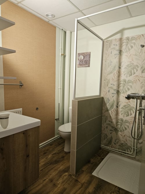 Salle de bains moderne aux Gîtes Les Muriers avec douche en carrelage beige, lavabo blanc, toilettes et papier peint botanique à Bourgvilain.