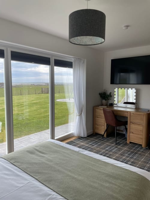 Modern bedroom at Flotnar Hus with sea view through glass doors, wooden desk, flat-screen TV, and garden views in Keiss