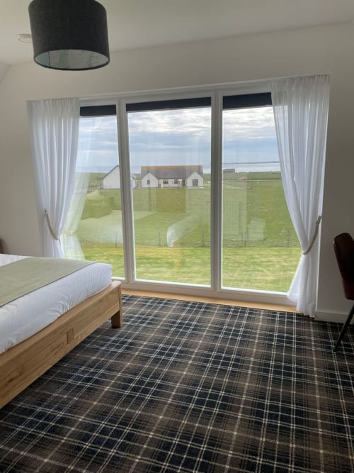 Modern bedroom with sea view at Flotnar Hus, featuring bed, plaid carpet, and large windows overlooking Scottish farmland and