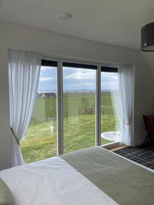 Bright bedroom with sea view through large glass doors, white sheer curtains, bed in foreground, green fields and coastal