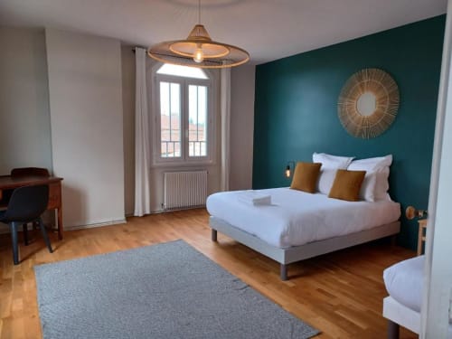 Chambre moderne avec mur d'accent bleu-vert, lit blanc avec coussins dorés, parquet, luminaire suspendu et vue sur le jardin