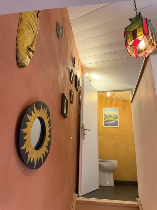 Flur, der in das obere Stockwerk des Ferienstudios „Occitan Heritage“ in La Sarrasine in Aiguèze führt, mit Terrakotta-Wänden, dekorativen Masken und einer handgefertigten Glaslampenleuchte, die zum Badezimmer führt.