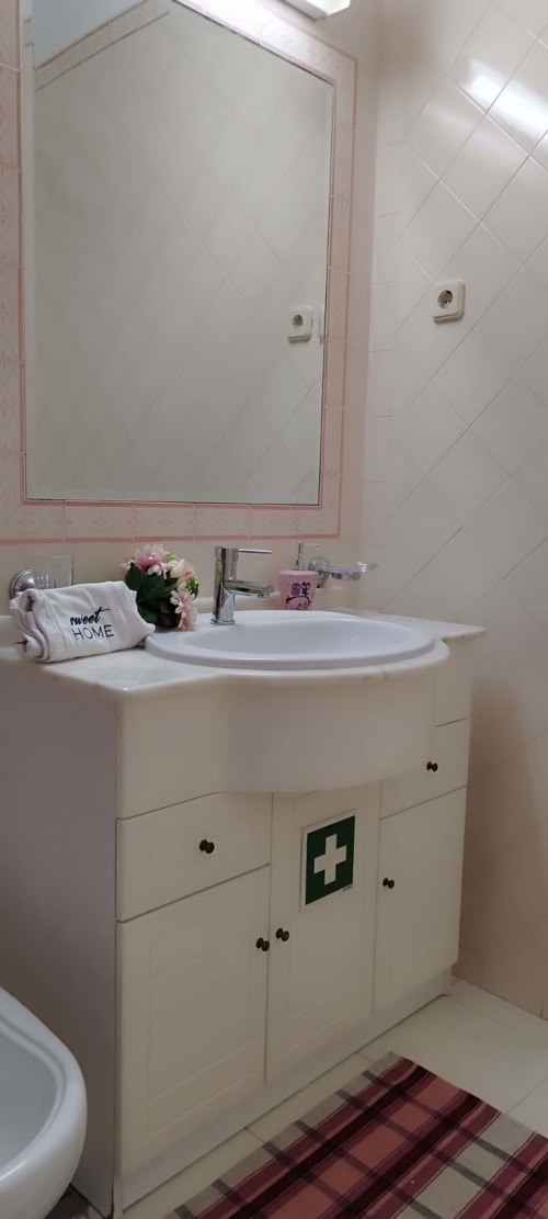 Bancada de banheiro moderno com pia redonda, espelho com moldura rosa, móveis brancos com cruz verde e toalha decorativa