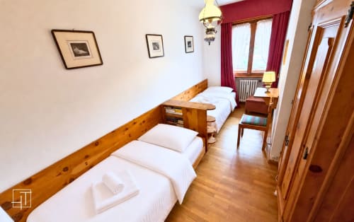 Twin bedroom with wooden beds, white linens, hardwood floor, mountain view windows with red curtains at Cortina d'Ampezzo