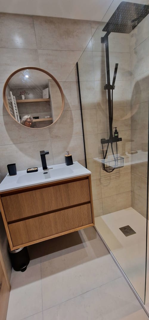 Salle de bain moderne du studio Zoppas Aix-les-Bains avec meuble lavabo en bois, miroir rond, robinetterie noire et douche à l'italienne