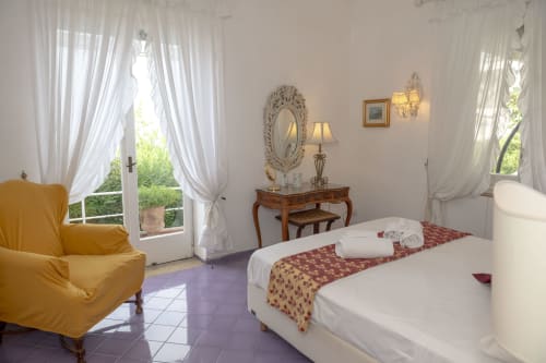 Camera luminosa a Villa La Capperina a Capri con letto bianco, poltrona gialla, specchio ornamentale e vista sul giardino