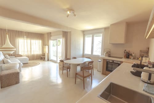 Modern open-plan hotel suite with kitchen, dining area, and bedroom featuring beige tones and bright natural light