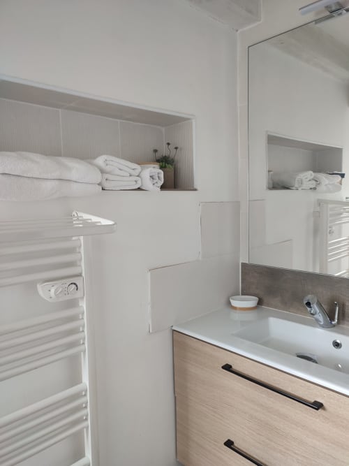 Salle de bain moderne aux murs blancs, meuble en bois, lavabo, miroir, sèche-serviettes chauffant et étagères avec serviettes blanches chez Orval Et