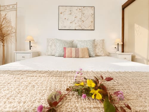 Dormitorio luminoso con ropa de cama blanca, almohadas florales y flores de jardín en primer plano en la Casa Celso de Baiona