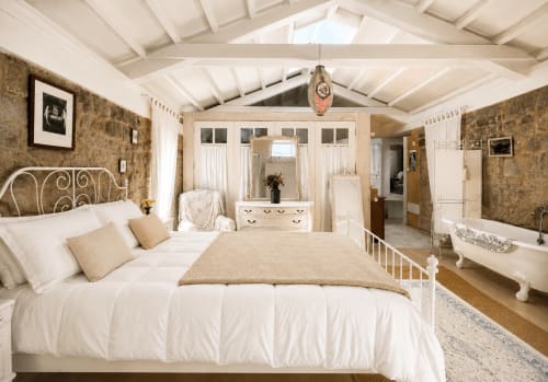 Dormitorio elegante en casita Casa Celso, Baiona, con cama de hierro blanco, muros de piedra vista y baño en suite vintage