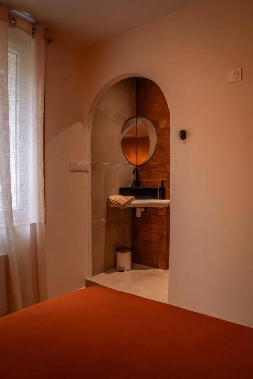 Salle de bains en terre cuite avec alcôve cintrée, miroir circulaire et vasque noire à la Maison Antea au Puy-en-Velay