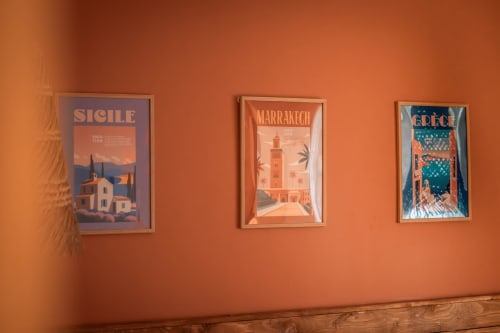Trois affiches de voyage vintage pour la Sicile, Marrakech et la Grèce affichées sur un mur orange dans la chambre La Parenthèse Solaire à