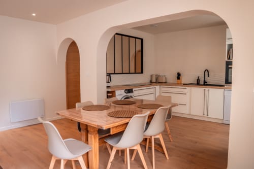 Coin repas et kitchenette du gîte Maison Antea au Puy-en-Velay avec table en bois, chaises blanches et cuisines crème