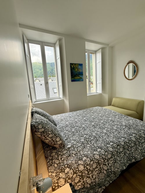 Bright bedroom with patterned bedding, mountain views from dual windows, decorative mirror, green chair at Les meublés