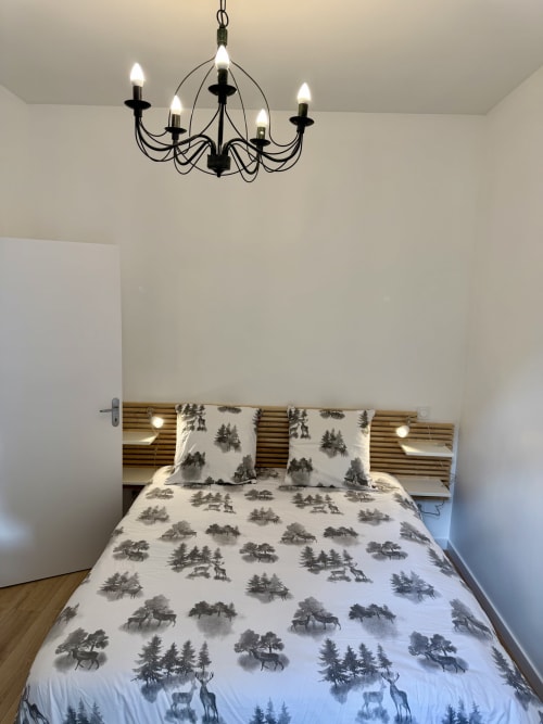 Modern bedroom with forest-patterned bedding, wooden headboard with built-in lights, and black chandelier at Les meublés
