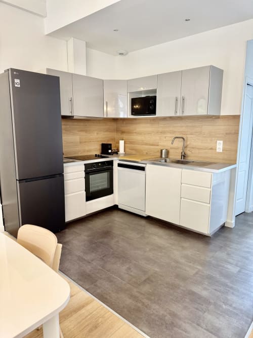 Modern L-shaped kitchen with white cabinets, wood backsplash, black appliances, and stainless steel sink at Les meublés