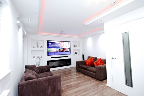 Modern living room with brown sofas, orange cushions, flat-screen TV, fireplace, and pink ceiling lights in Salford apartment