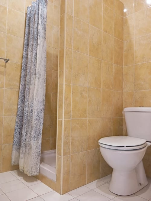 Casa de banho com chuveiro azulejado em bege, sanita branca e cortina estampada no apartamento Sweet Home, Portimão