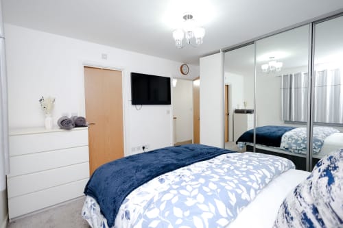 Modern bedroom at ShelterHomes UK in Salford with blue bedding, wall-mounted TV, mirrored wardrobe, and white furnishings