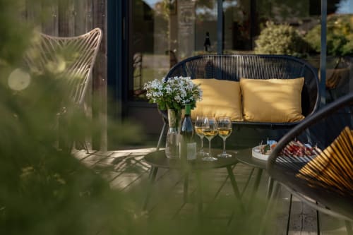 Élégante terrasse du Manoir de la Nogrie avec mobilier en rotin noir, fleurs blanches, verres de vin et coussins jaunes