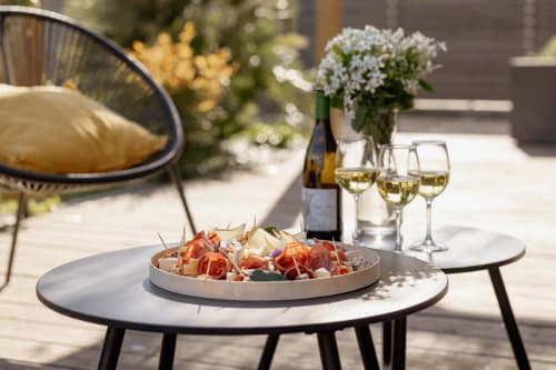 Terrasse ensoleillée du Manoir de la Nogrie avec amuse-bouches, vin blanc, fleurs et mobilier contemporain à Val d'Izé, France